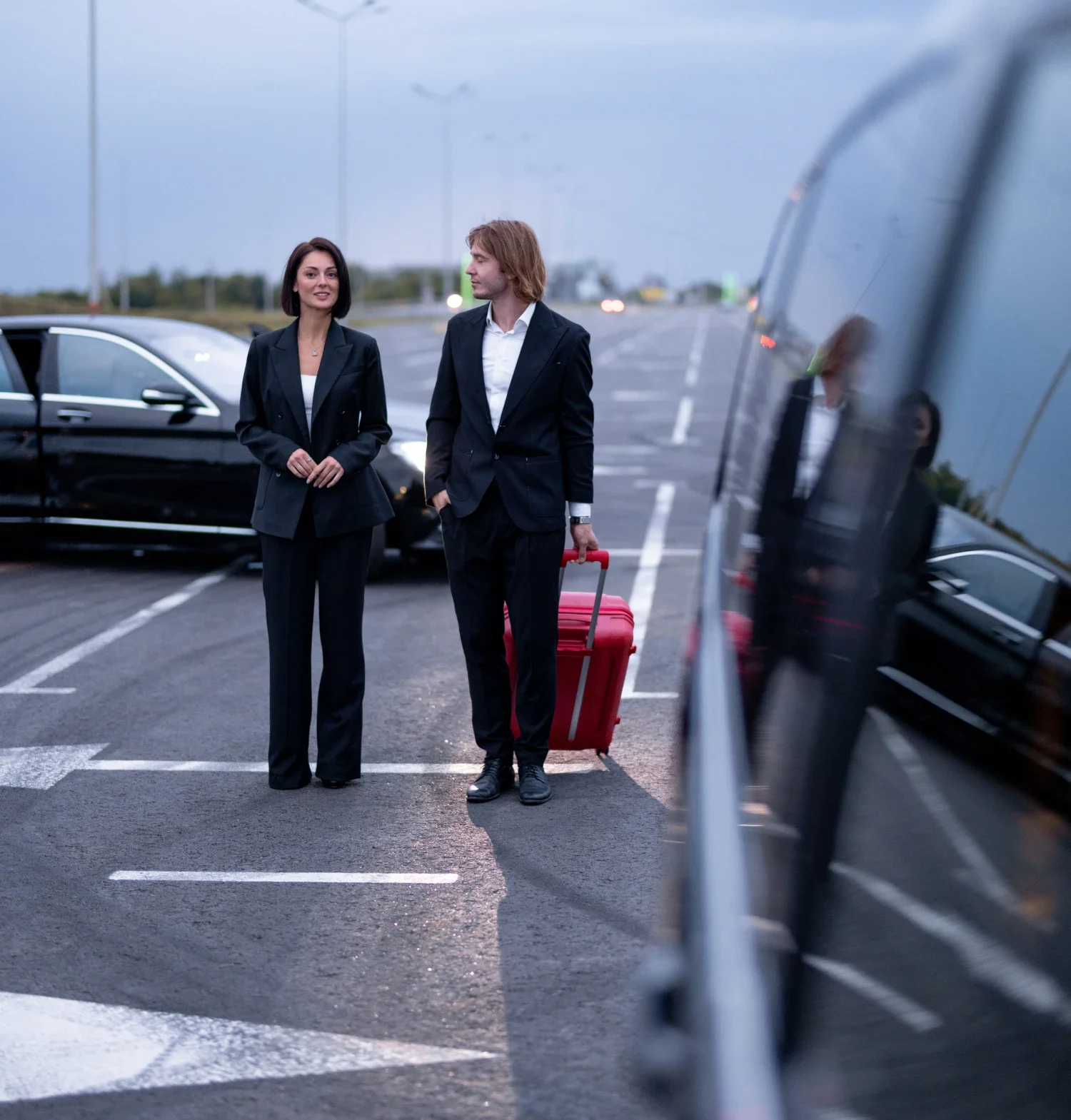 Two business professionals stand by a car, engaged in outdoor conversation.